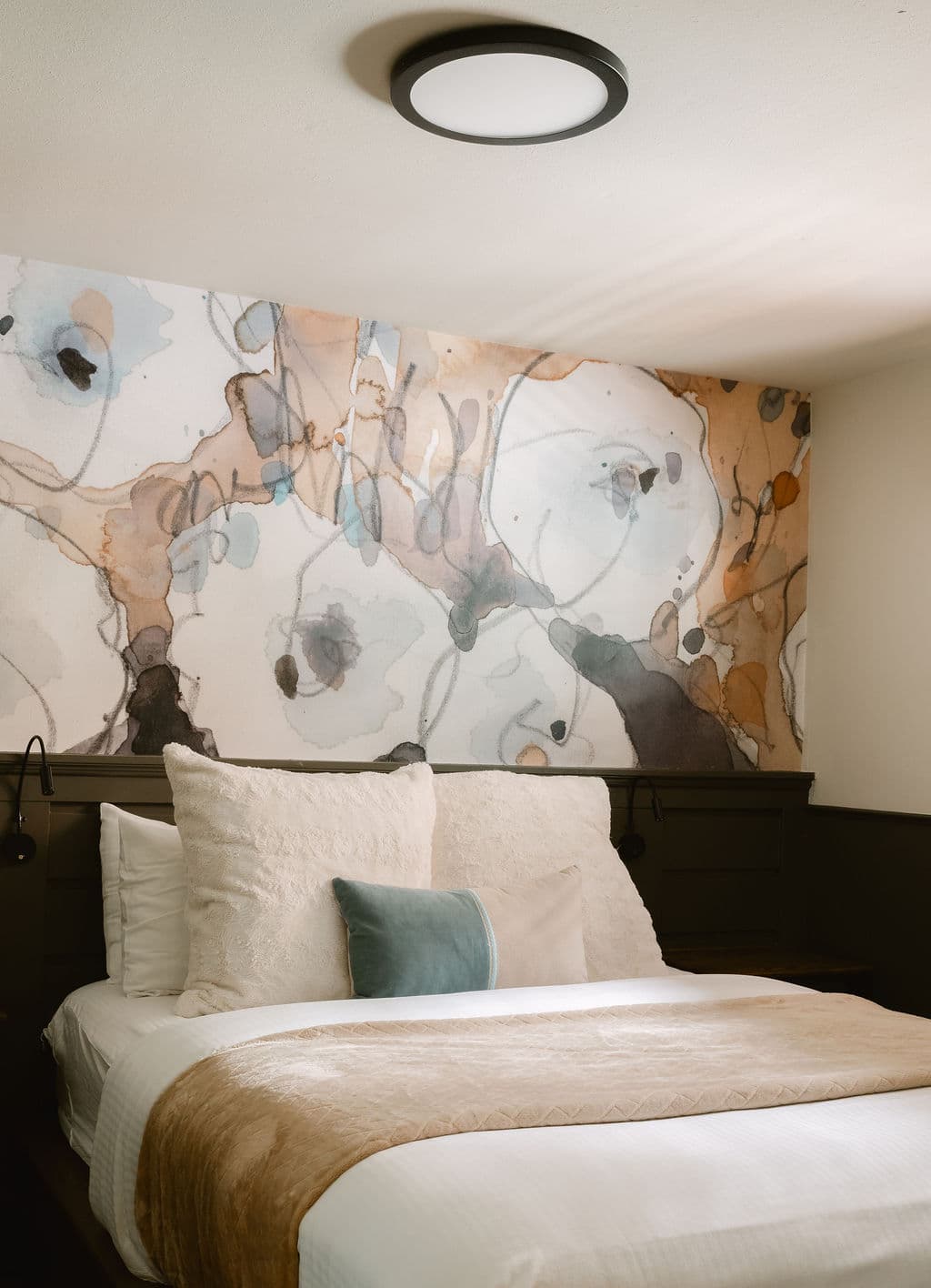 A cozy bedroom features a bed with beige and white linens, plush pillows, and a blue cushion. There's an abstract floral mural above and soft lighting, creating a serene atmosphere.