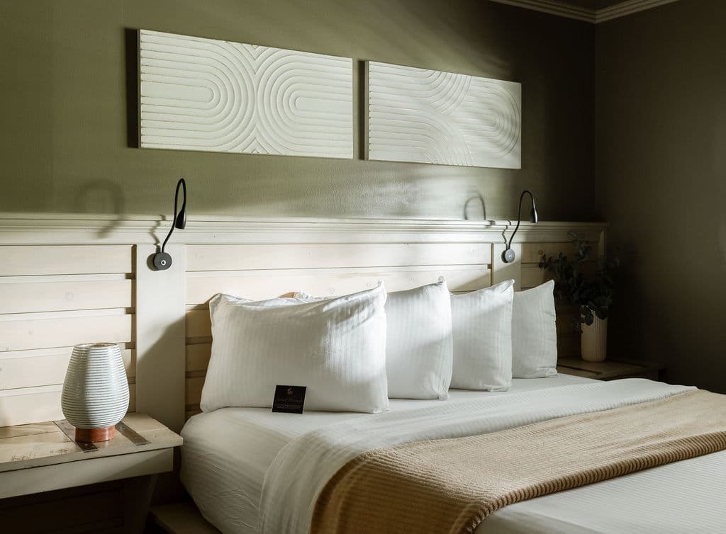 A cozy bedroom with a white bedspread, several pillows, modern wall art, and warm lighting.