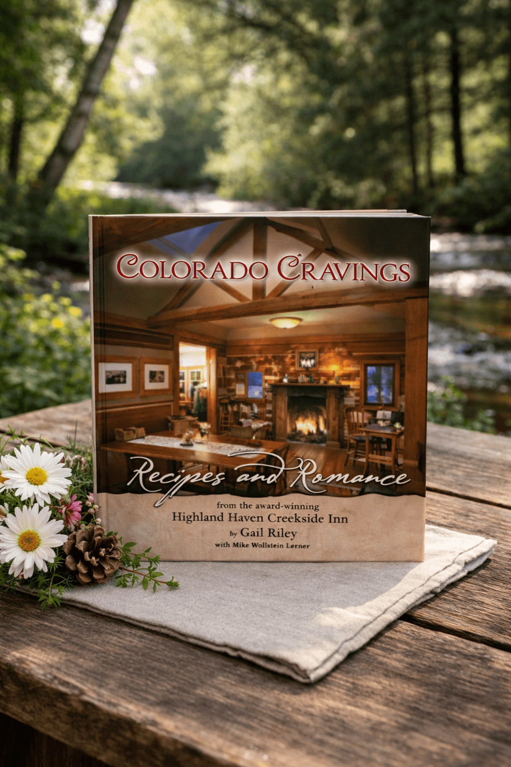 A cookbook titled "Colorado Cravings: Recipes and Romance" sits on a wooden table beside flowers and a creek in a forest setting.