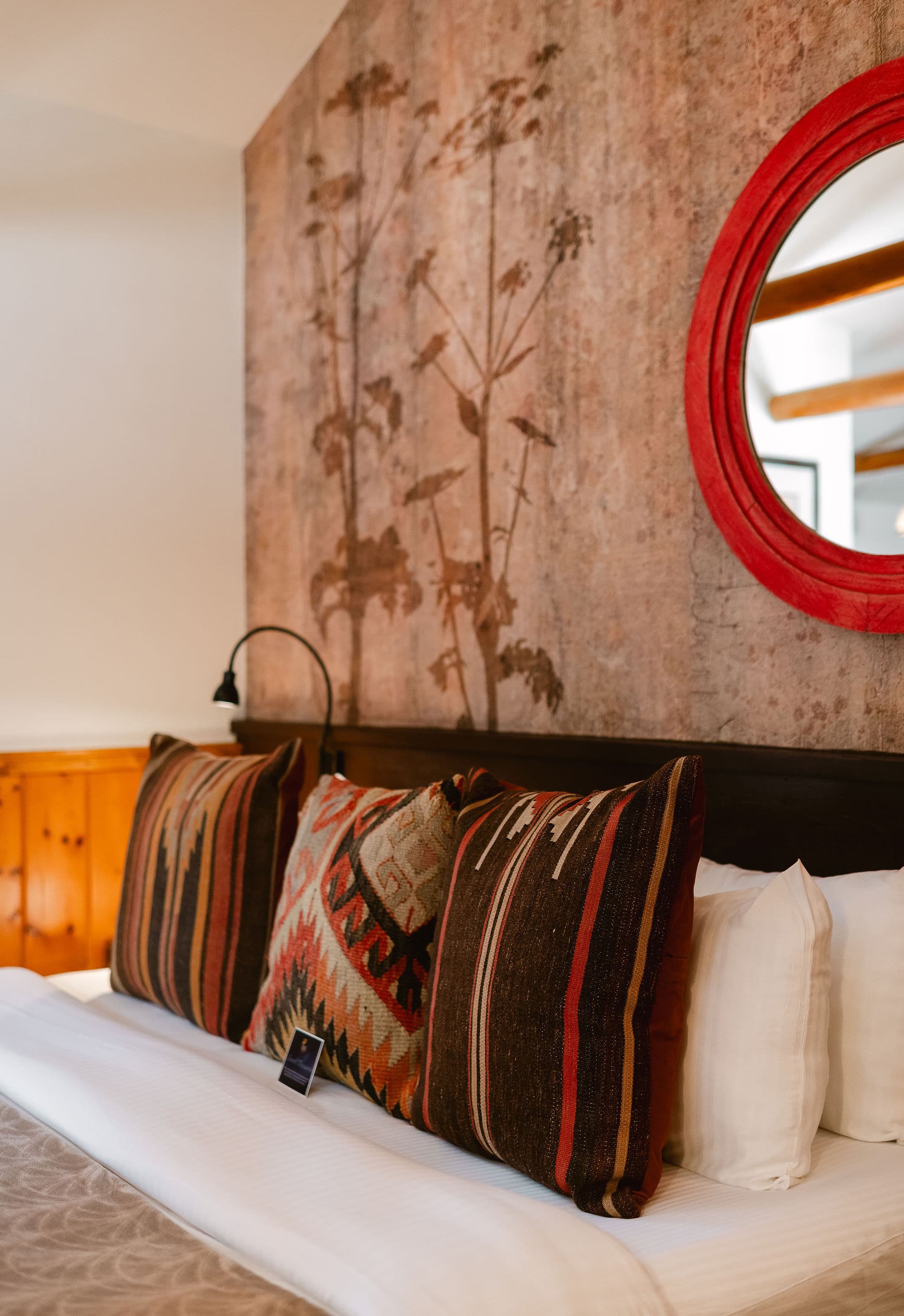 A cozy bed with patterned pillows against a textured wall and a circular mirror.