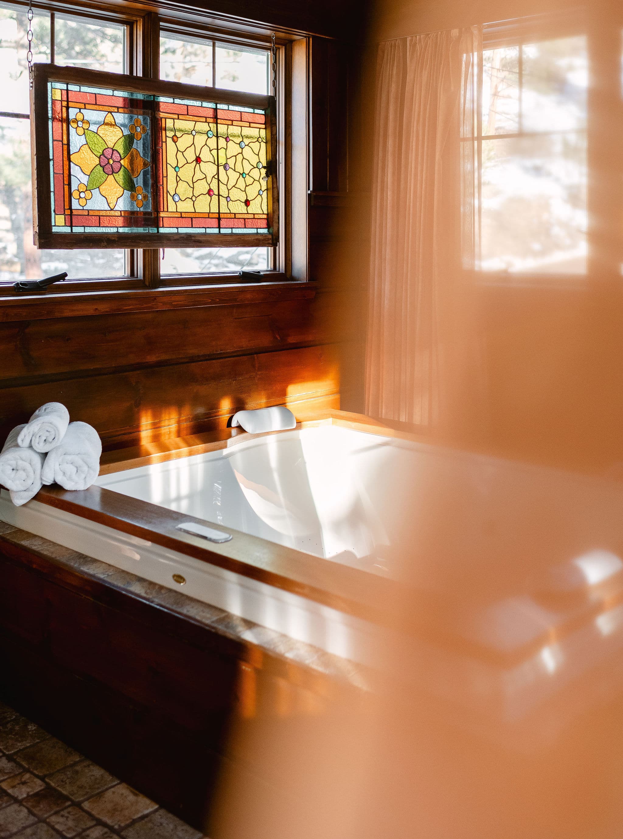 A well-lit bathroom with a wooden hot tub and neatly rolled towels beside it, framed by a colorful stained glass window.