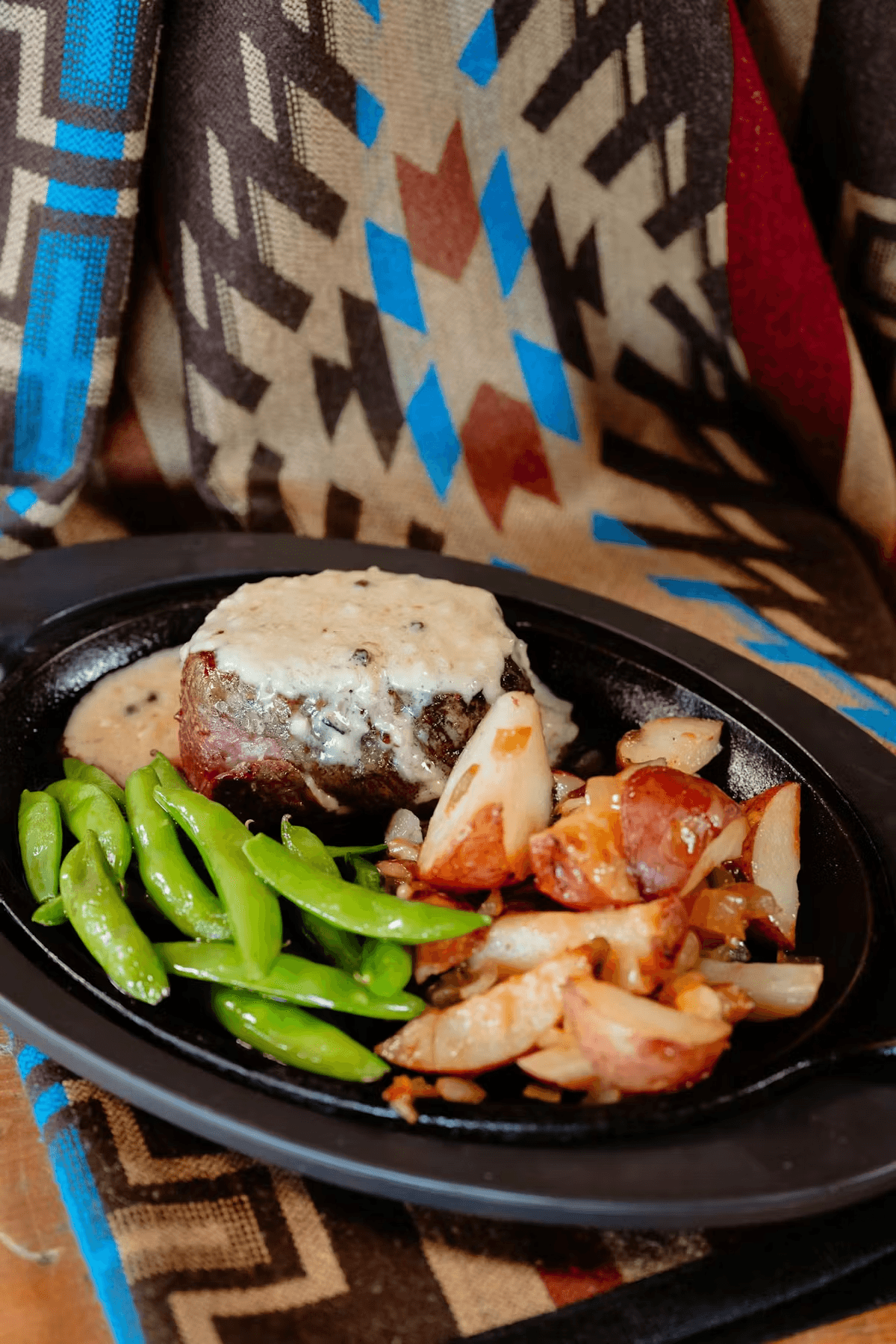 A plate featuring a beef steak with creamy sauce, green beans, and roasted red potatoes, set against a patterned textile background.