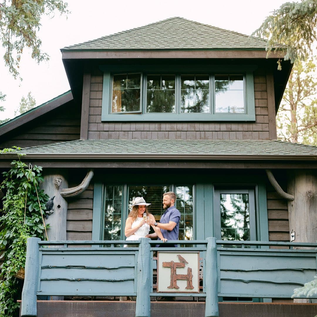 A couple stands on the porch of a rustic cabin surrounded by trees. The woman wears a hat, sipping from a glass, while the man smiles warmly beside her.