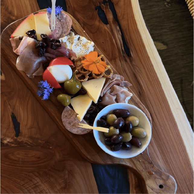 A wooden board displays an assortment of meats, cheeses, olives, fruit, and garnishes.