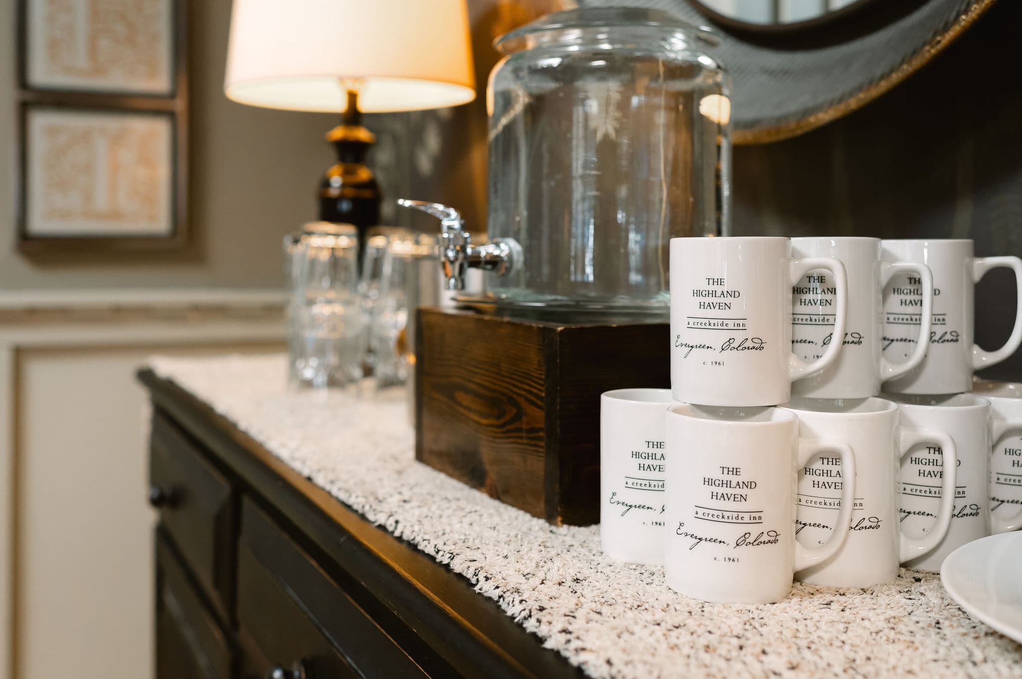 A wooden table displays branded coffee mugs and a glass water dispenser beside a lamp.