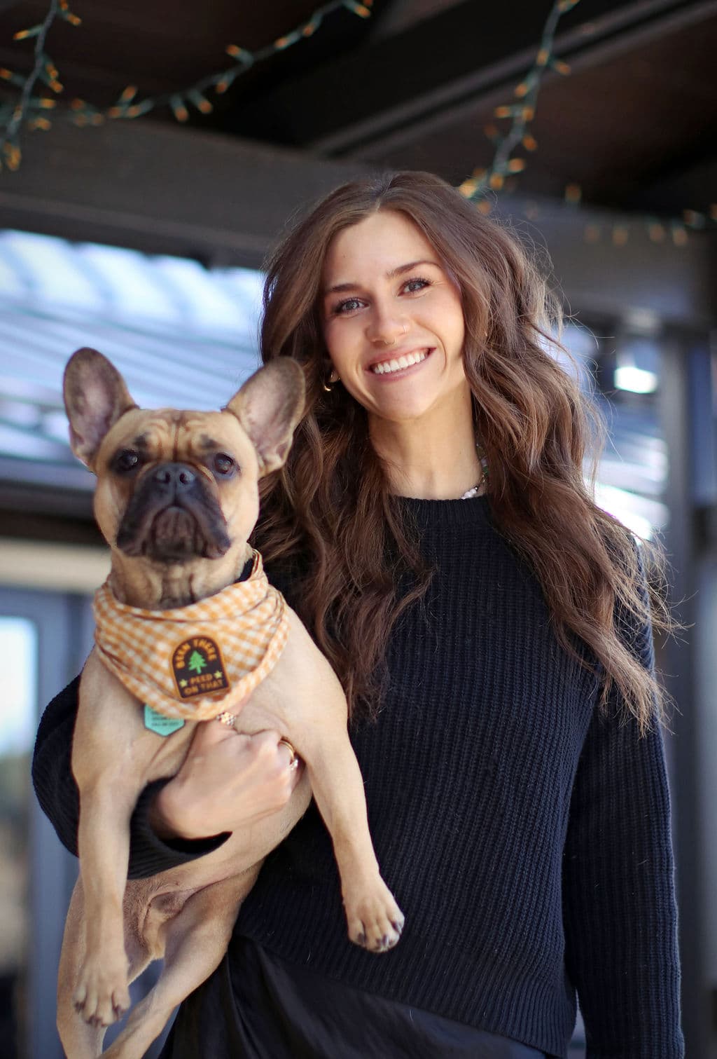 A smiling woman holds a French Bulldog while standing outdoors.