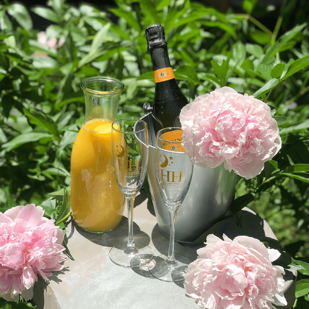 A bottle of champagne, a carafe of orange juice, two flutes, and pink peonies arranged on a table surrounded by greenery.