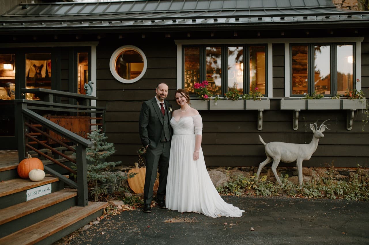 A couple stands in front of a rustic building, smiling, with a decorative deer statue nearby.