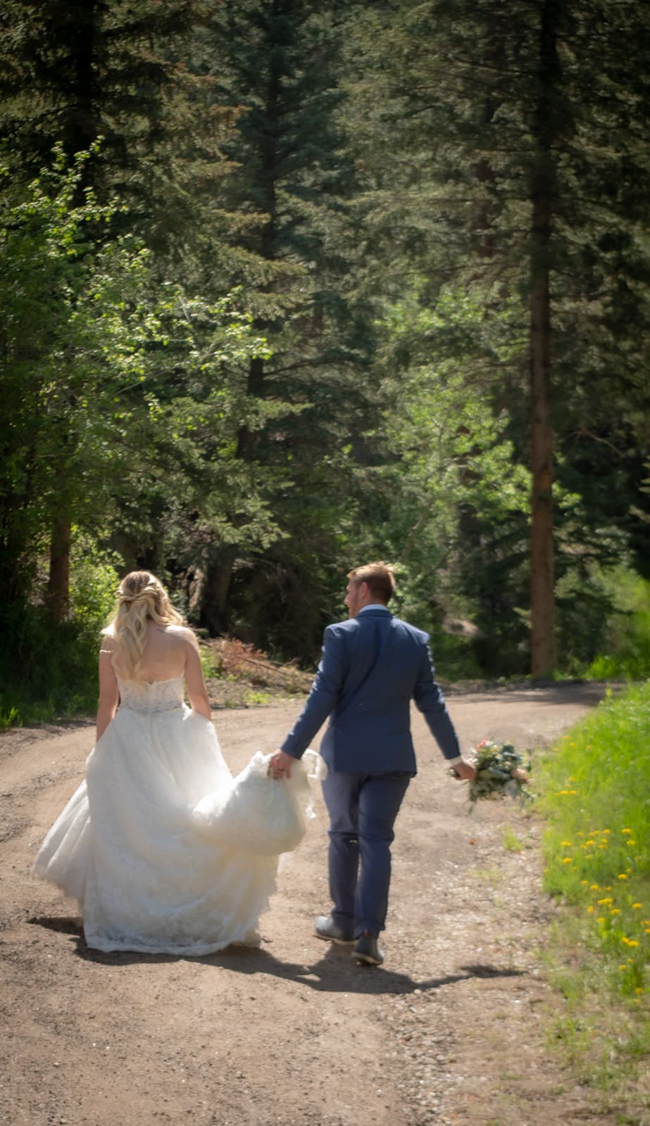 A bride and groom walk hand in hand along a dirt path surrounded by trees.