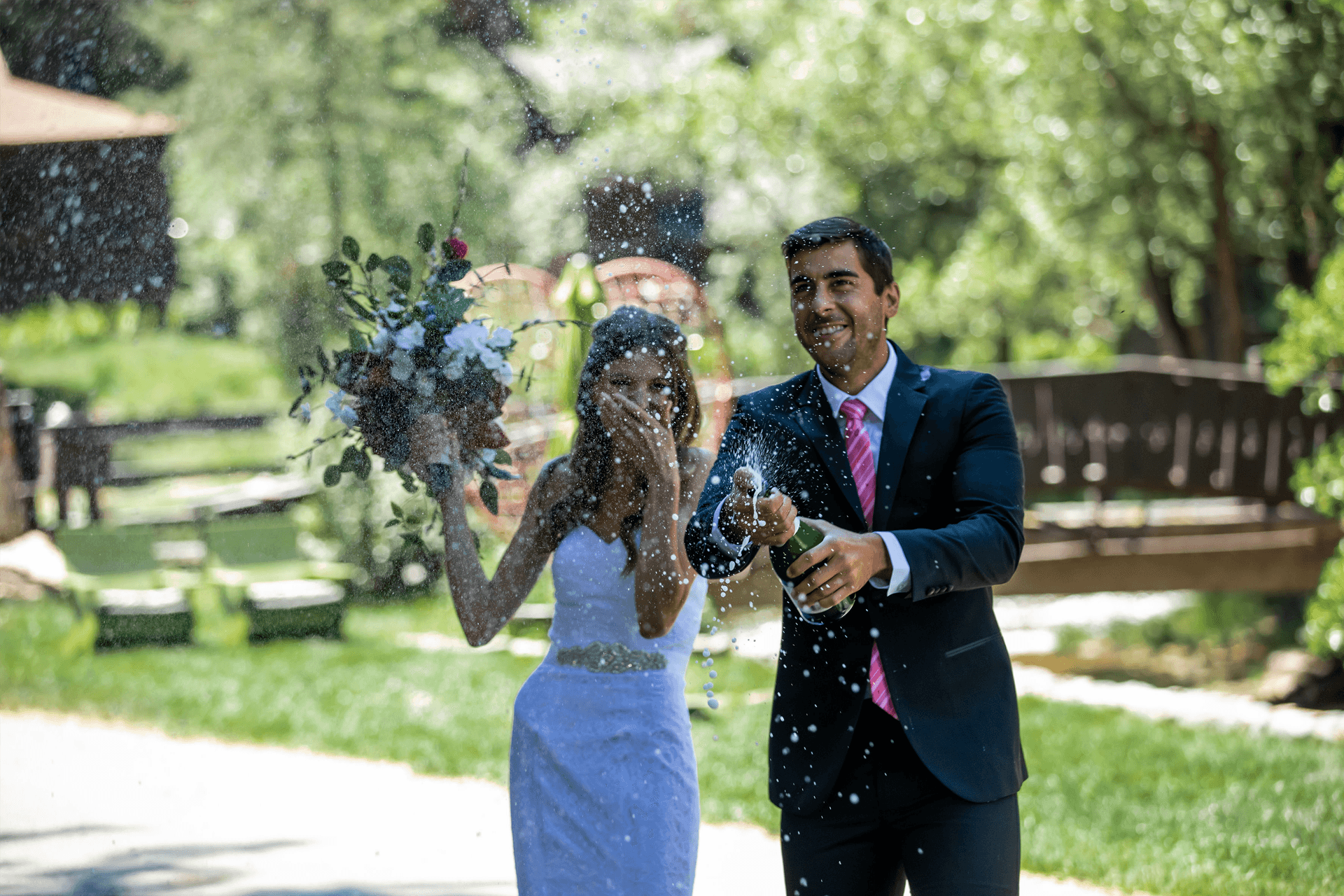 A smiling couple celebrates with champagne while the bride holds a bouquet, surrounded by lush greenery.