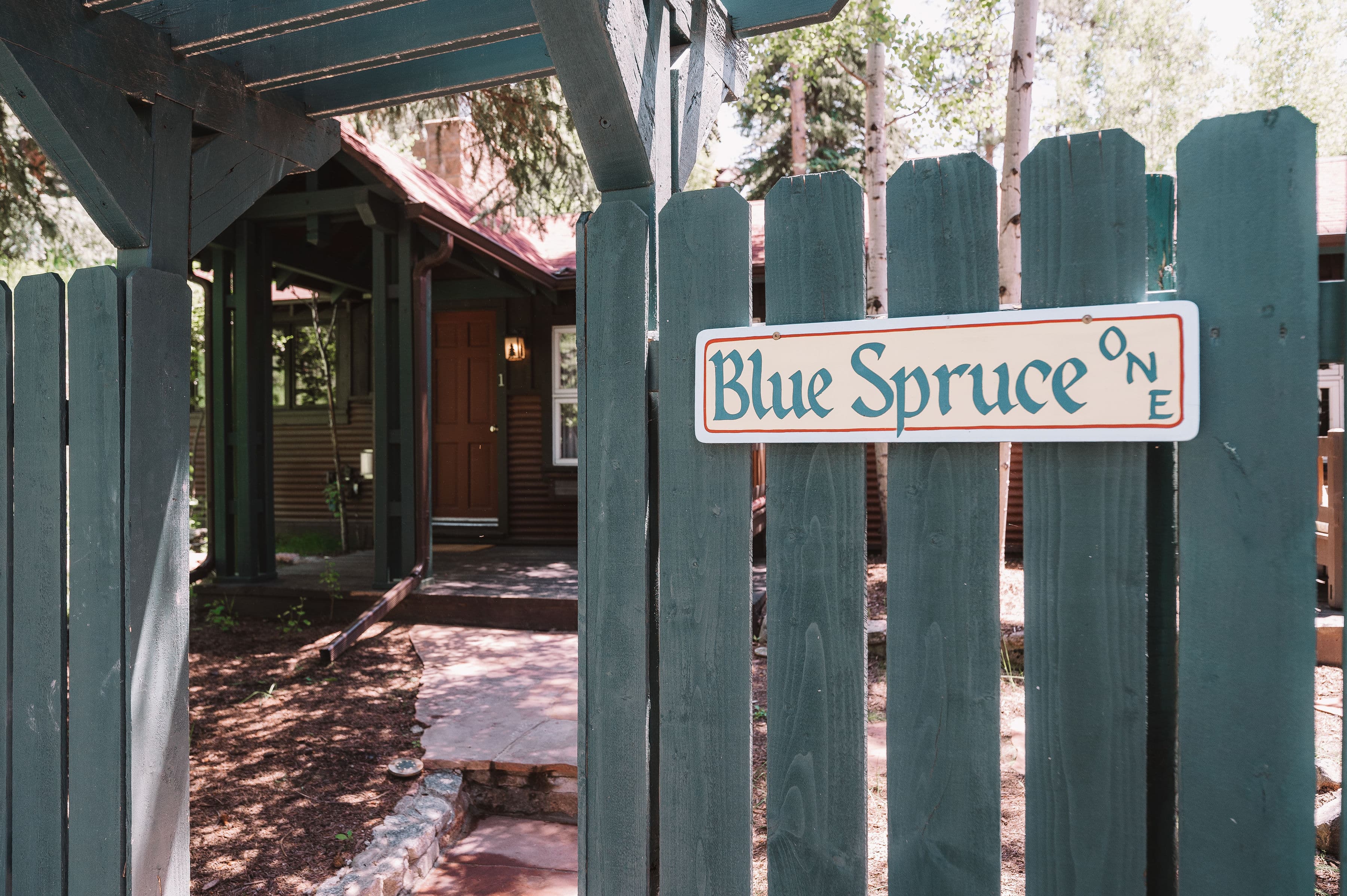 A wooden fence with a sign reading "Blue Spruce One" leads to a cozy cabin.