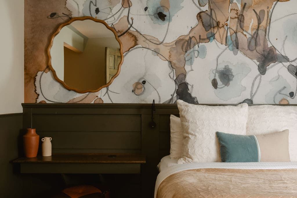 Bedroom with abstract watercolor-patterned wallpaper and a scalloped round mirror. Dark wood headboard, cozy white pillows, and neutral bedding add warmth.