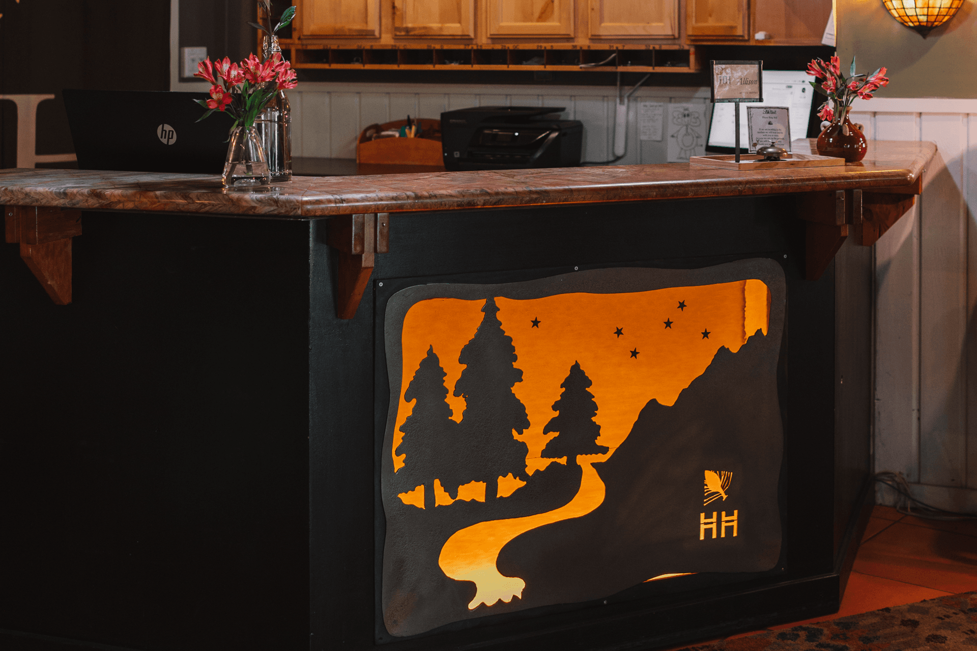 A wooden reception desk with floral decor. The front panel features a glowing silhouette of trees and a river under starry skies, creating a warm ambiance.