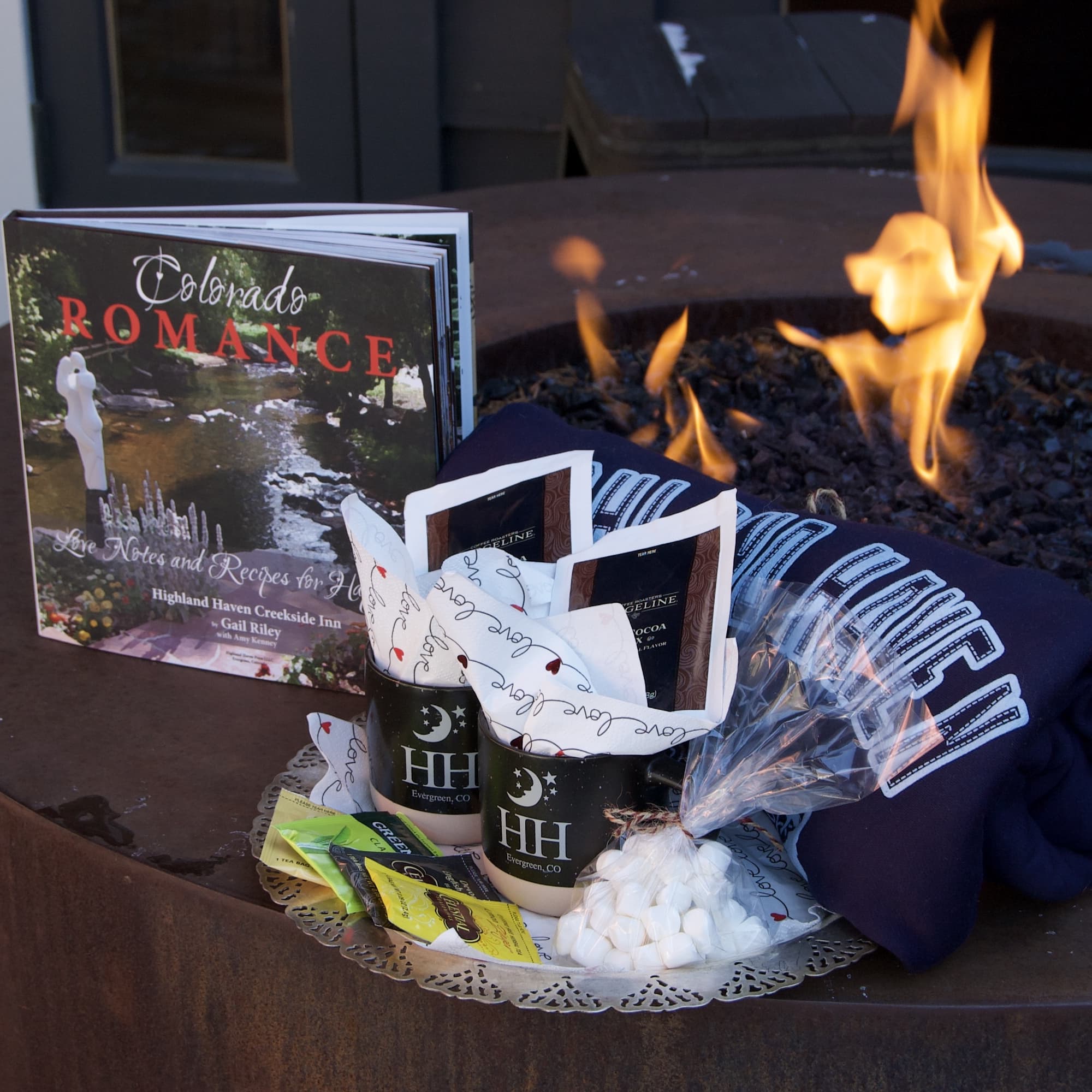 Cozy outdoor setup featuring a book, hot chocolate supplies, marshmallows, a hoodie, and a fire pit.