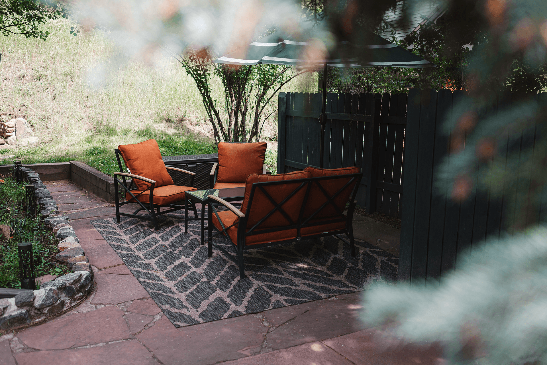 Outdoor patio with cozy seating, featuring orange cushions and a patterned rug. Surrounded by greenery, trees, and a wooden fence, conveying a serene atmosphere.