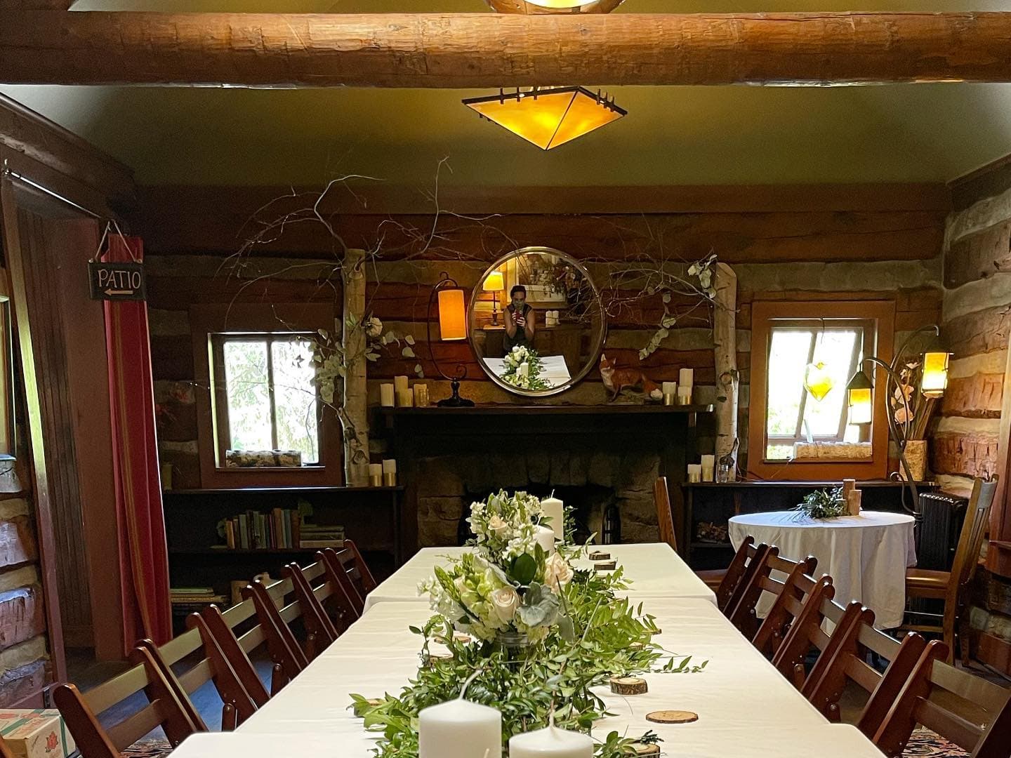 A warmly lit rustic dining room with a long table adorned with greenery and candles, featuring a stone fireplace and wooden decor.