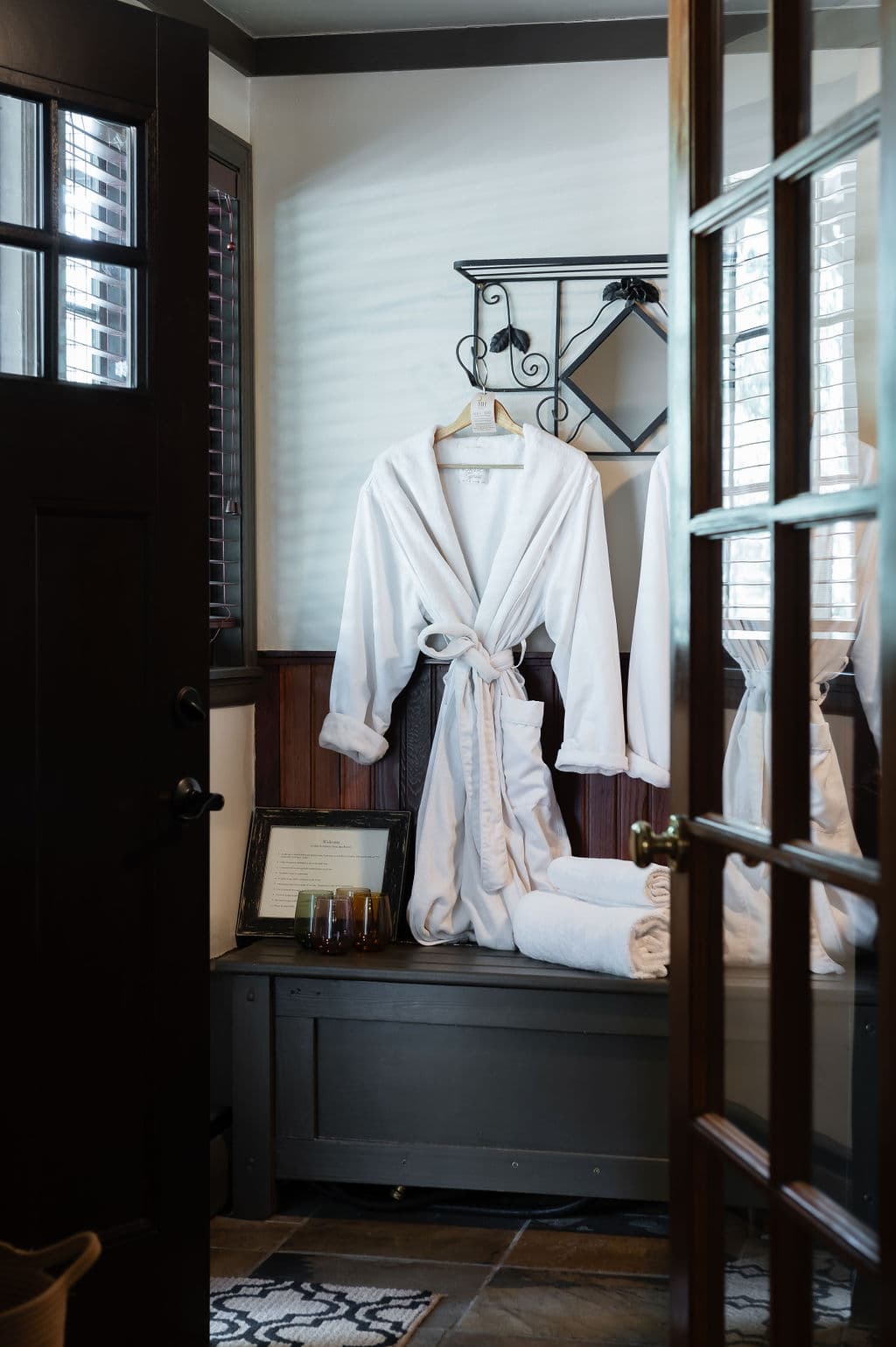 A white bathrobe hangs on a hook with neatly stacked towels and a framed menu nearby.