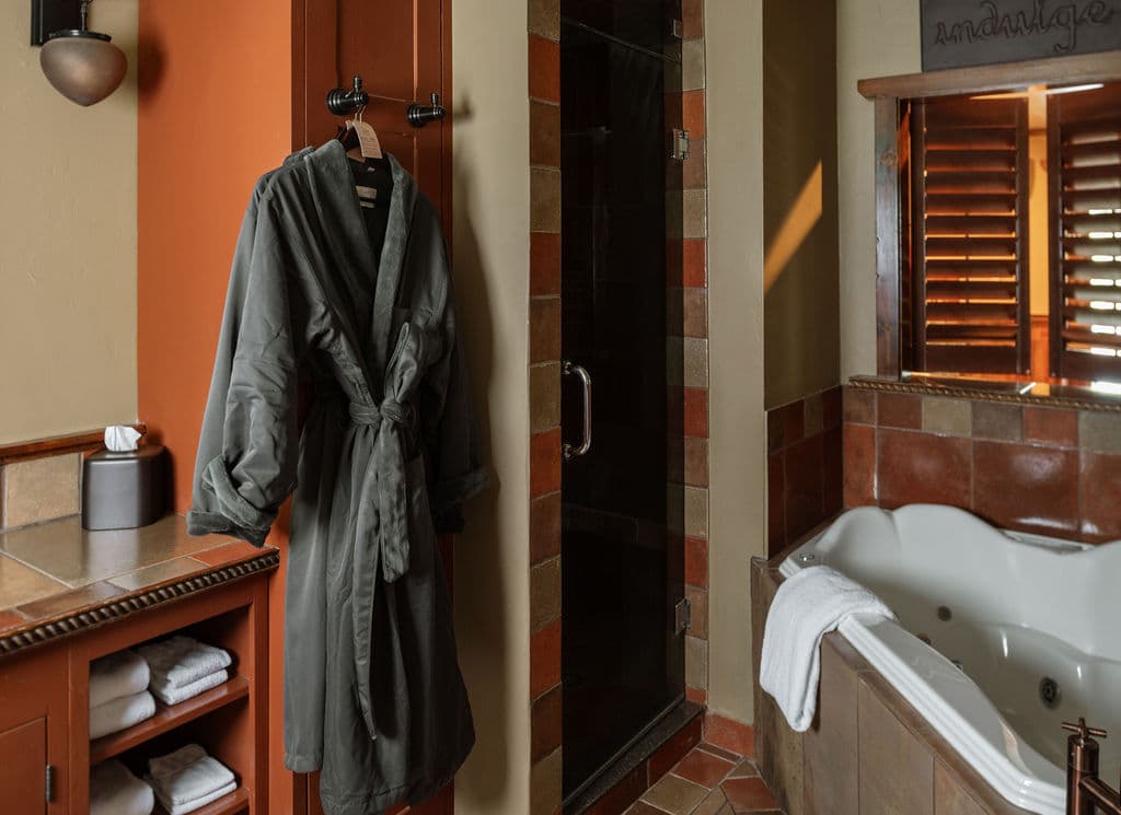 A plush gray bathrobe hangs in a stylish bathroom featuring a shower, a jacuzzi, and neatly stacked towels.