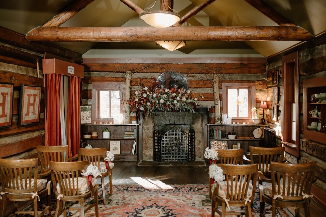 A cozy rustic room with wooden beams, arranged seating, and a floral display above a stone fireplace.