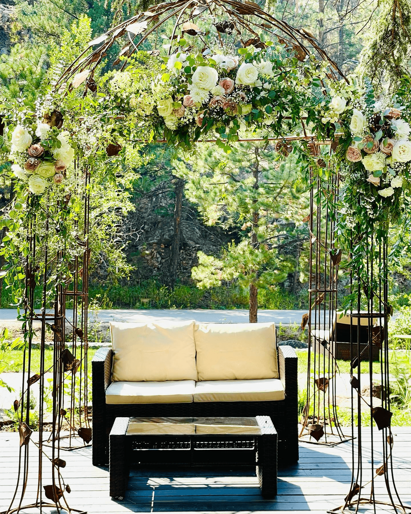 A cozy outdoor seating area framed by a floral arch and surrounded by greenery.