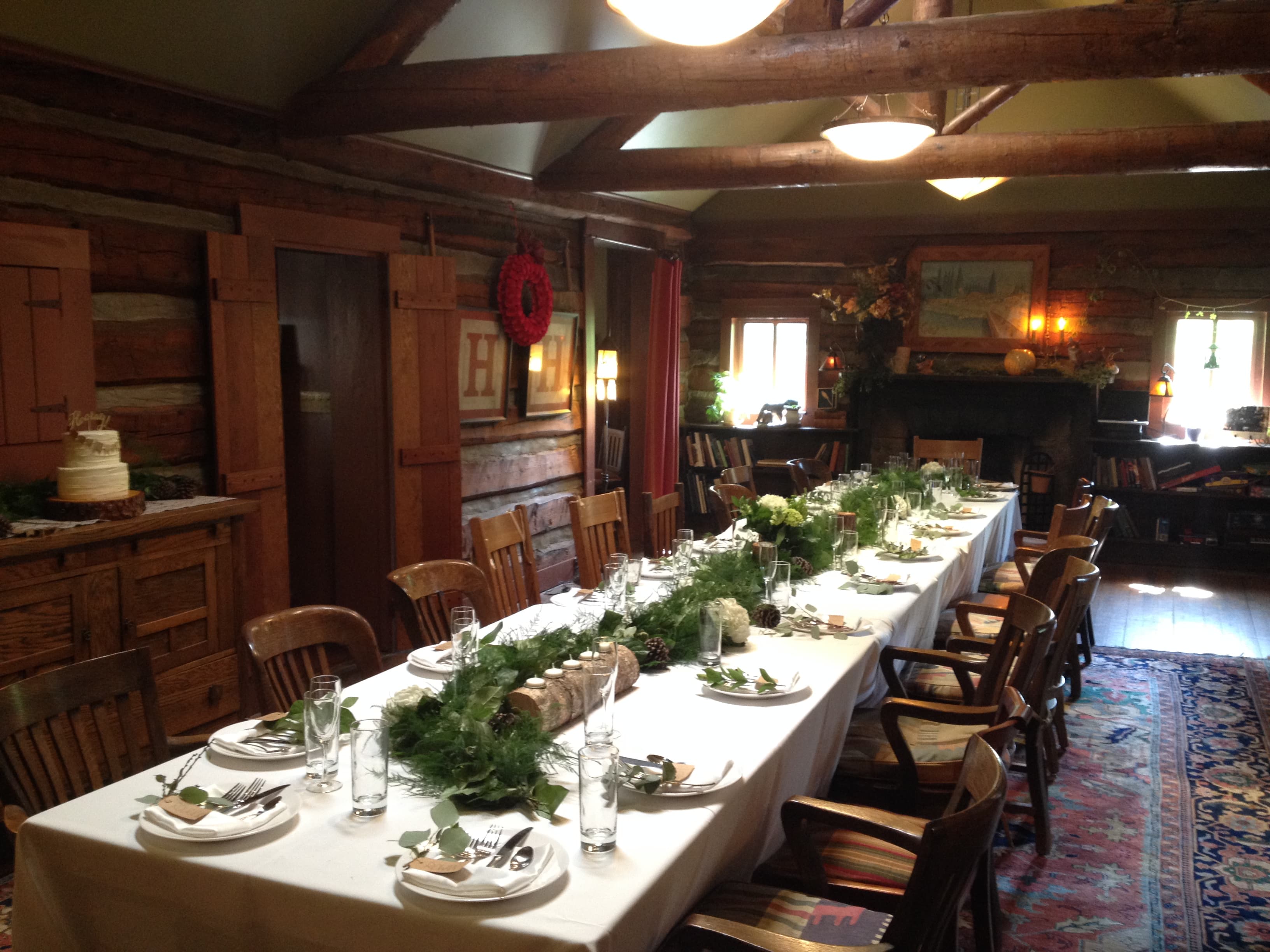 A rustic dining room with a long table set for a meal, featuring greenery, elegant table settings, and wooden accents.