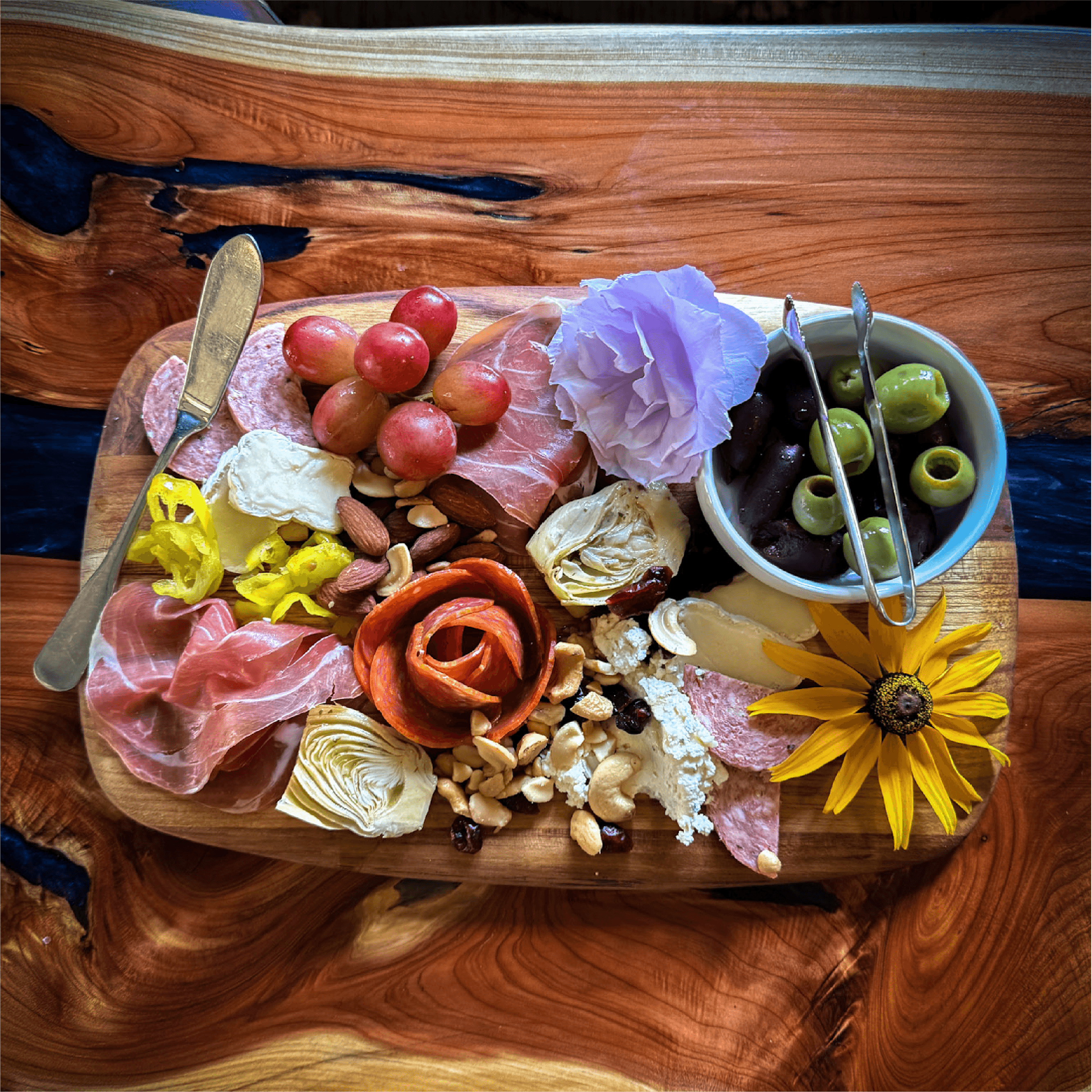 A wooden serving board filled with assorted meats, cheese, fruits, nuts, and decorative flowers.