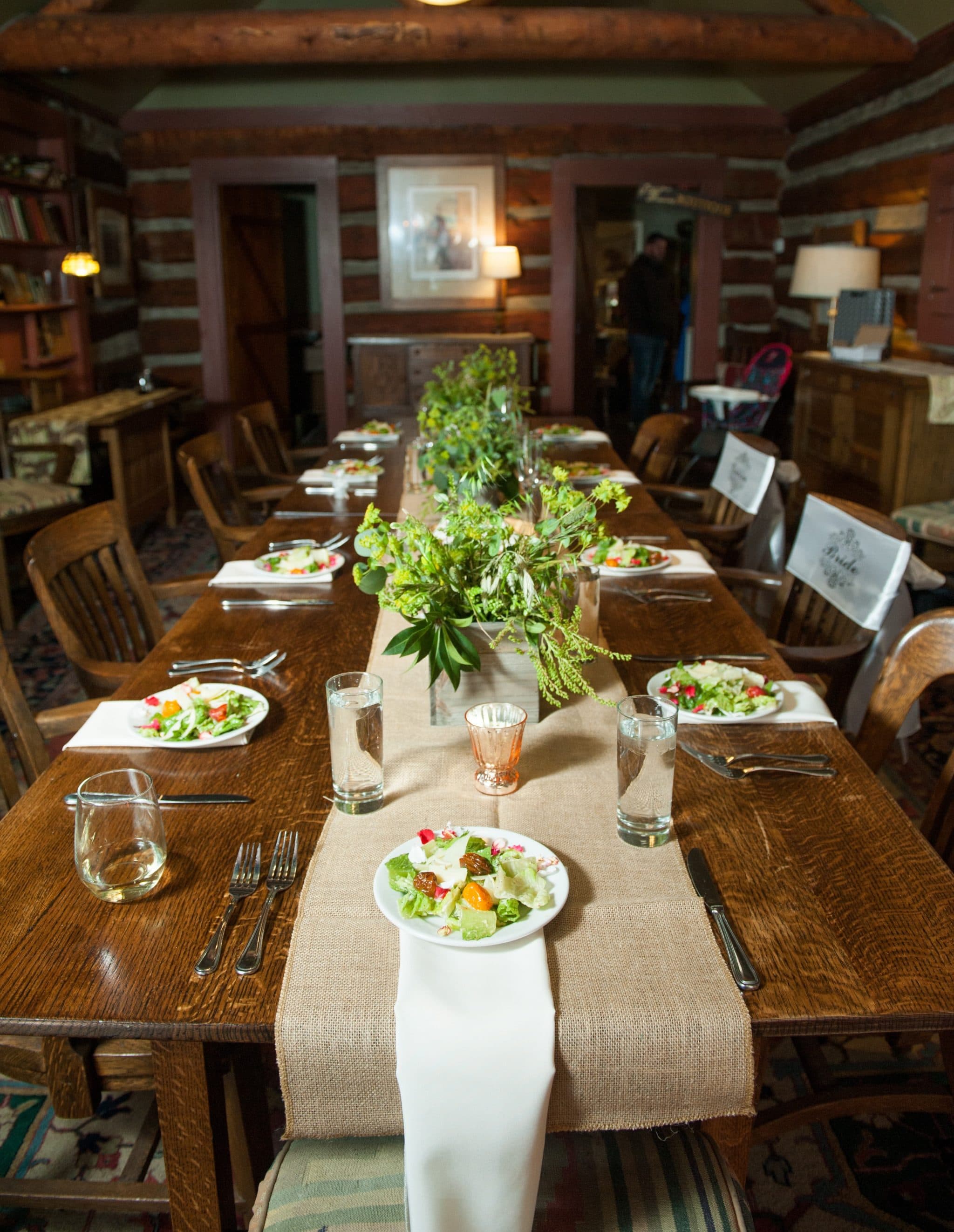 A long dining table is set with plates of salad, glasses of water, and a floral centerpiece in a rustic room.