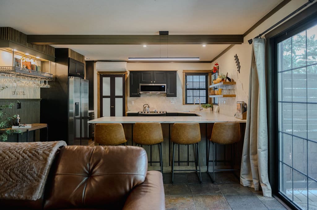 Modern kitchen with a warm atmosphere, featuring a marble island with four brown leather bar stools. Large window on right brings in natural light.