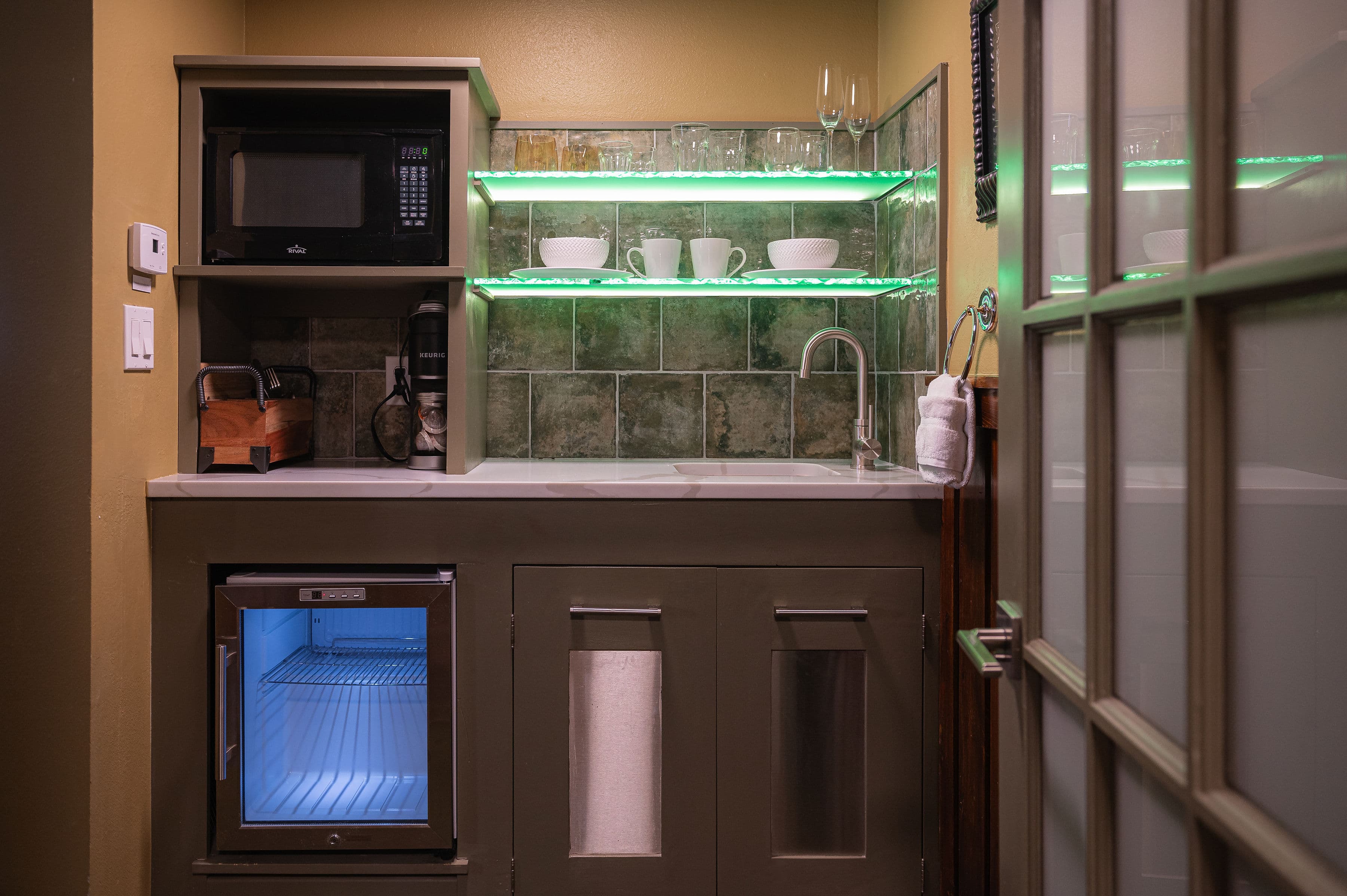 A compact kitchenette featuring a microwave, mini fridge, and glassware on illuminated shelves.