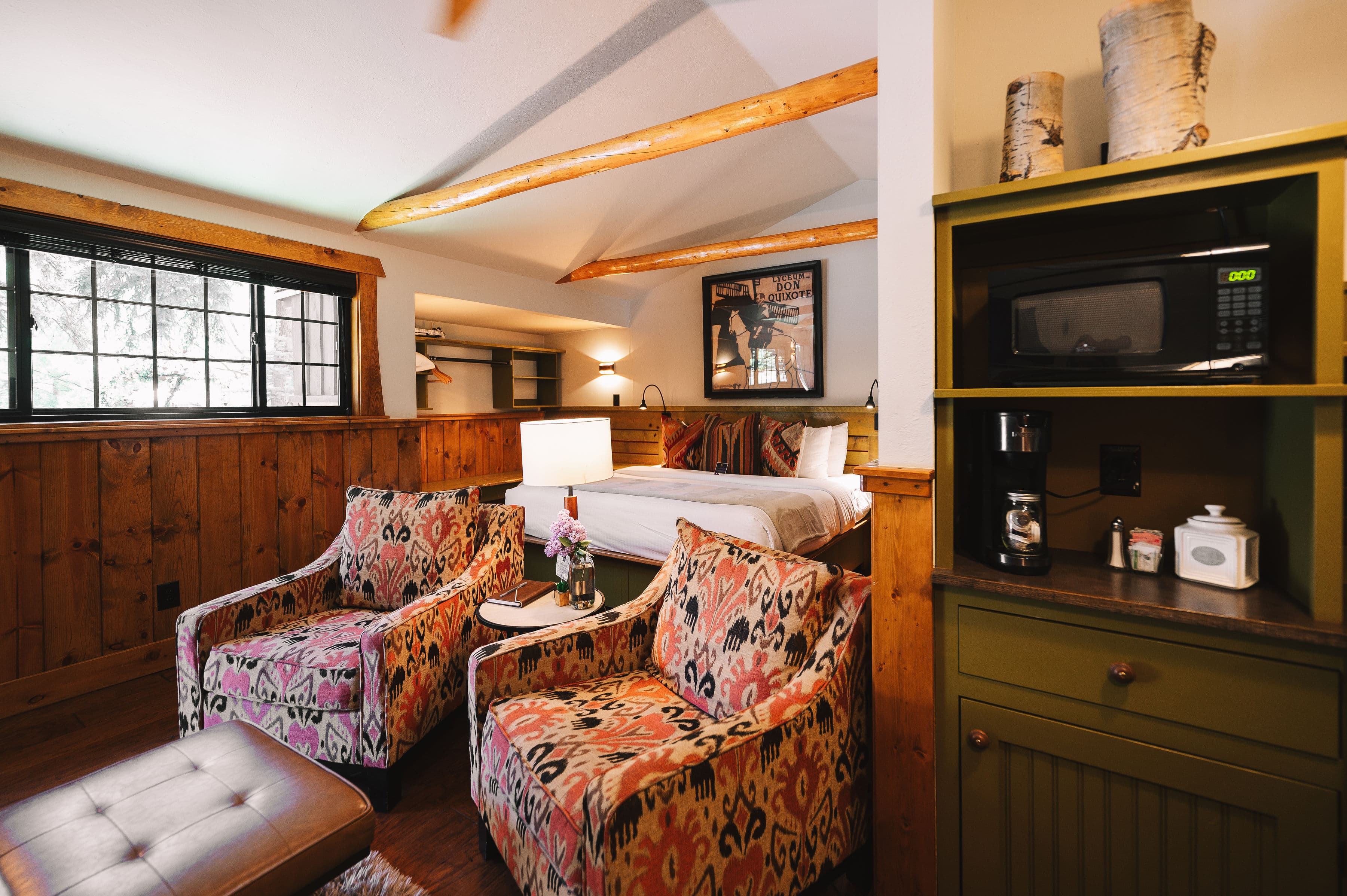 Cozy rustic room with exposed wooden beams, a bed, patterned armchairs, a coffee table with flowers, large window, and a kitchenette with a microwave.