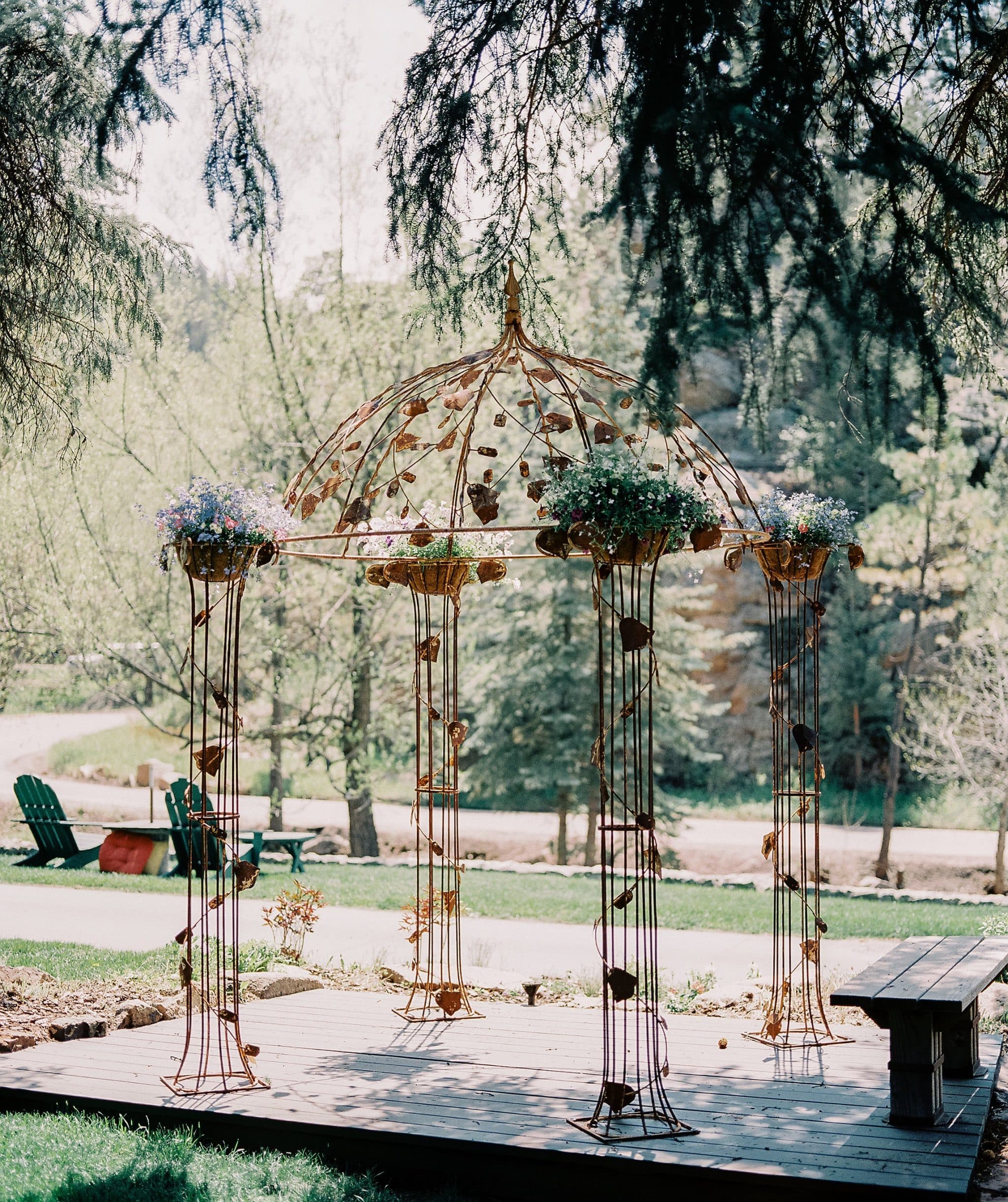 A decorative metal gazebo adorned with potted flowers on a wooden deck in a lush garden setting.
