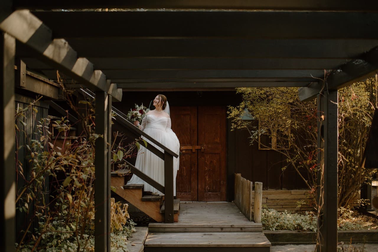 A bride in a flowing white gown ascends wooden steps, holding a bouquet, framed by rustic architecture and autumn foliage.
